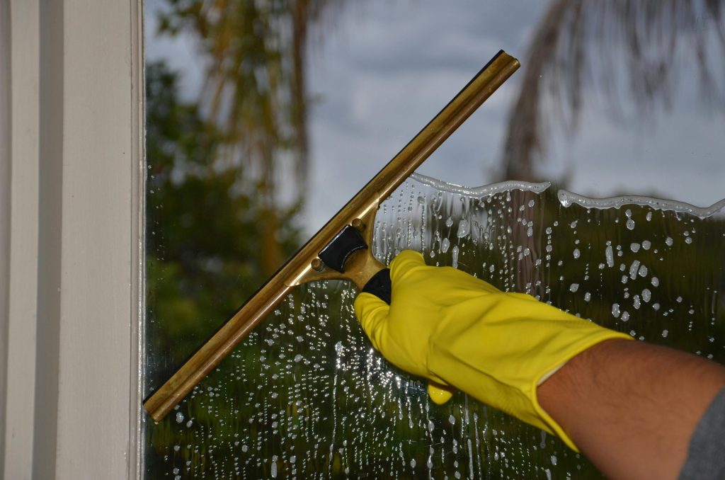 Nahaufnahme einer Hand, die im Freien mit einem Abzieher und gelben Handschuhen ein Fenster reinigt.